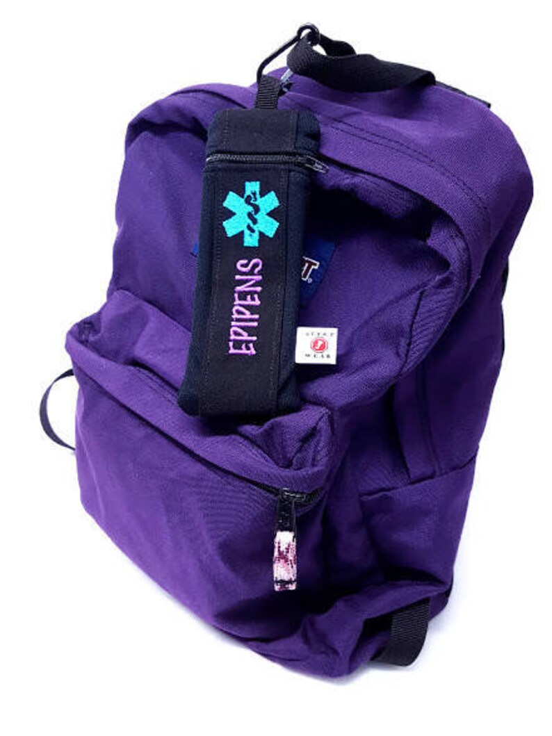 May include: Purple backpack with a black zippered pouch attached to the front. The pouch has a blue and green star of life symbol and the text "EPIPENS" printed on it.