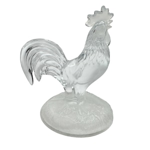 May include: A clear glass rooster figurine. The rooster is standing on a frosted, circular base. The details of the rooster's feathers and comb are visible. The figurine is a decorative item.