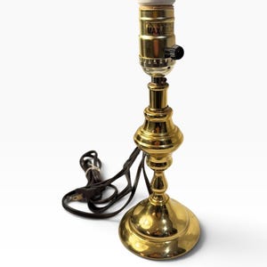 May include: A polished brass table lamp base with a rounded pedestal and a central column. The socket features a black turn-knob switch and text reading CAUTION RISK OF FIRE MAX 60W TYPE A LAMP. A brown electrical cord is attached to the base.