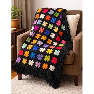 Könnte beinhalten: Eine schwarze Häkeldecke mit bunten quadratischen Mustern in verschiedenen Farben, darunter Rot, Orange, Gelb, Grün, Blau und Lila. Die Decke hat einen schwarzen Fransenbesatz und liegt über einem braunen Sessel.