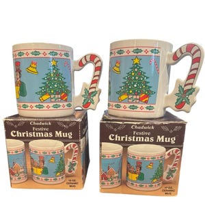 May include: Two white ceramic Christmas mugs with candy cane handles. Each mug features a Christmas tree, gifts, and a bell on a light blue background. The mugs are on top of boxes that say "Chadwick Festive Christmas Mug".