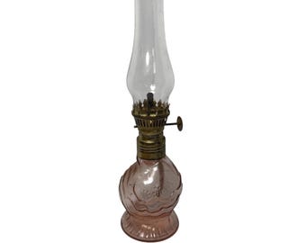 May include: A vintage-style oil lamp featuring a clear glass chimney, a brass-toned metal burner assembly with an adjustment knob, and a rounded, pale pink glass base with a molded, scalloped, and embossed pattern throughout the surface.