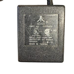 May include: A black Atari power supply unit with the Atari logo and text including "PART NO. C010472" and "MADE IN TAIWAN". It has UL and SP certification marks.