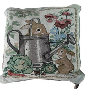 May include: A square throw pillow featuring a woven tapestry design of two brown rabbits in a garden setting. One rabbit sits inside a grey metal watering can, while the other stands beside it. The scene includes green cabbage, carrots, and flowers.