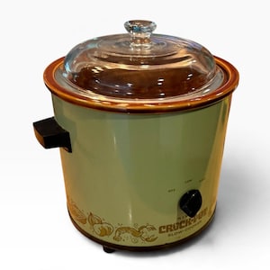 以下が含まれることがあります： 薄緑色のCrock-Potスロークッカー。茶色の縁と透明なガラス蓋が付いています。本体にはオフ、低、高の設定ができる黒いコントロールノブがあり、底部には野菜の装飾的なグラフィックがプリントされています。