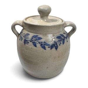 May include: A beige stoneware crock with a matching lid and two side handles. The exterior features a blue painted vine pattern encircling the upper portion, along with a small rectangular stamped mark near the rim of the vessel.