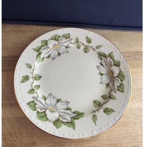 May include: White ceramic plate with a floral design. The plate features three large white flowers with green leaves and vines around the rim. The edge of the plate has a gold trim.