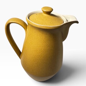 May include: A mustard yellow ceramic teapot with a speckled finish, featuring a matching lid with a small knob, a curved handle on the side, and a wide pouring spout. The interior of the teapot and the rim are a contrasting off-white color.