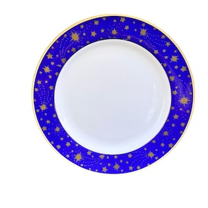 May include: A round dinner plate with a plain white center and a wide cobalt blue rim. The rim features a repeating pattern of gold-colored stars and radiating starbursts of varying sizes, finished with a thin gold-colored trim along the edge.