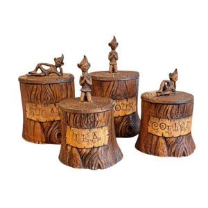 May include: Set of four brown ceramic canisters with elf figurines on the lids. Each canister is shaped like a tree stump and labeled "SUGAR", "TEA", "FLOUR", and "COFFEE". The canisters have a rustic, vintage aesthetic.