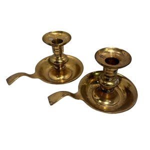 May include: A pair of polished brass chamberstick candle holders, each featuring a circular drip tray, a central stem with a flared top, and a curved handle extending from the side. The metal surface shows a warm, reflective golden tone.