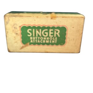 May include: A vintage Singer buttonhole attachment box. The box is rectangular with a cream-coloured exterior and a green top. The front features a green and red label with the Singer logo and "Buttonhole Attachment" text.