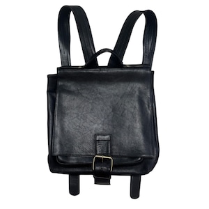 May include: A black leather backpack featuring a large front flap with a single brass-toned metal buckle closure. The bag includes a top handle and two adjustable shoulder straps, showcasing a textured surface and a structured rectangular shape.