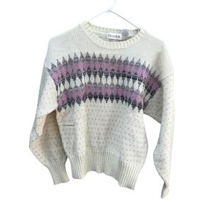 May include: Cream-colored sweater with a geometric pattern in shades of pink, gray, and black across the chest. The sweater has a ribbed crew neck, cuffs, and hem. Small, evenly spaced dots cover the body of the sweater.