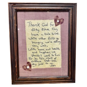 May include: A dark brown wooden frame containing a beige fabric panel with black embroidered text that reads: Thank God for dirty dishes, they have a tale to tell. While other folks go hungry, we're eating very well. With home and health and happiness, we shouldn't want to fuss. For by this stack of evidence, God's been very good to us. 1897. Two plaid fabric hearts with white buttons are attached.
