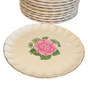 May include: A stack of cream-colored ceramic plates with scalloped edges and gold trim. Each plate features a central illustration of a vibrant pink camellia flower with green leaves and a small bud, set against a smooth, off-white background.