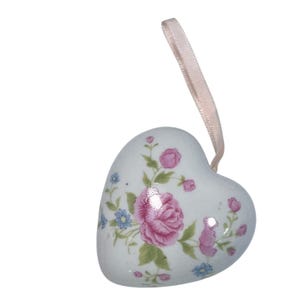 May include: A heart-shaped ceramic ornament featuring a white base with a floral pattern of pink roses, small blue flowers, and green leaves. A pale pink ribbon is attached to the top for hanging, providing a smooth and glossy finish.