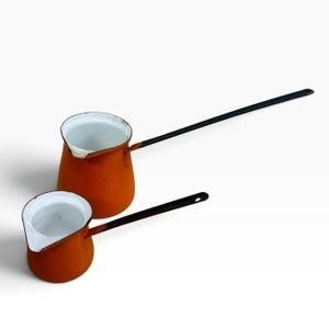 May include: Two orange enameled metal coffee pots with white interiors and long black handles. The pots feature a cylindrical body with a pouring spout on the rim. The handles are thin and straight, extending outward from the side of the pots.
