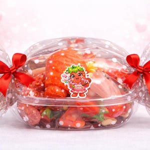 May include: A clear plastic container filled with sweets, wrapped in polka-dot cellophane and tied with red satin bows. A sticker of a strawberry character is on the front. The sweets are a mix of red, orange, and pink.