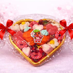Puede incluir: Una caja en forma de corazón llena de caramelos variados, envuelta en celofán transparente y atada con lazos de satén rojo. Los caramelos incluyen golosinas en forma de corazón, flor y redondas en tonos rosa, rojo y blanco. Un personaje de fresa de dibujos animados está en el centro.