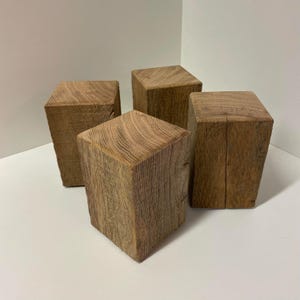 Op de afbeelding: Vier rechthoekige houten blokken gemaakt van natuurlijk, onafgewerkt hout. Elk blok heeft een duidelijk, zichtbaar houtnerfpatroon met een lichtbruine, verweerde textuur. De blokken zijn in een groep gerangschikt en tonen hun solide vorm.