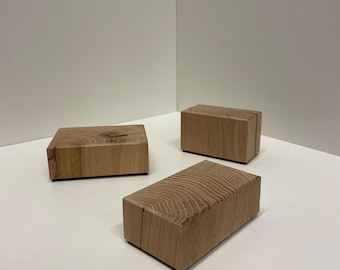 May include: Three rectangular blocks made of light brown natural wood. Each block displays a distinct grain pattern on its top surface, showing circular growth rings and wood texture. The blocks are arranged in a cluster, showcasing the raw wood.