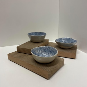 Könnte beinhalten: Drei Keramikschalen mit einer hellcremefarbenen Außenseite und einer blau gemusterten Innenseite. Jede Schale weist ein einzigartiges blaues Design mit Blumen- und geometrischen Motiven auf und steht auf einzelnen rechteckigen Holzblöcken.