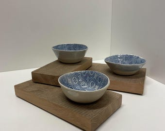 Könnte beinhalten: Drei Keramikschalen mit einer hellcremefarbenen Außenseite und einer blau gemusterten Innenseite. Jede Schale weist ein einzigartiges blaues Design mit Blumen- und geometrischen Motiven auf und steht auf einzelnen rechteckigen Holzblöcken.