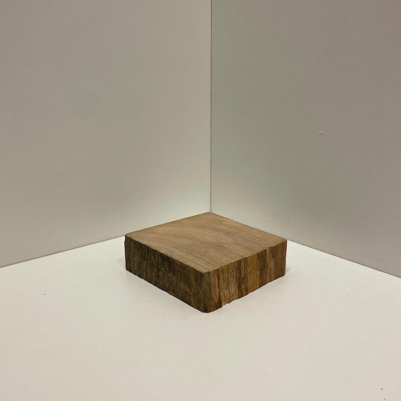 May include: A square wooden block with a natural grain pattern and rough, textured bark edges. The piece features a flat top surface and a thick, solid construction, showcasing the organic brown tones and wood grain texture of the material.