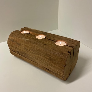 May include: A rustic, wooden candle holder with three lit tea light candles. The candle holder is made from a section of a weathered log, showcasing natural wood grain and texture. The warm glow of the candles contrasts with the brown wood.