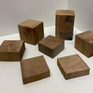 May include: A collection of seven natural wooden blocks of varying heights and square shapes. Each block displays a distinct light brown wood grain pattern, visible growth rings on the top surfaces, and rough, textured sides with natural cracks.