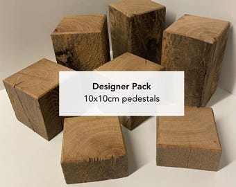 Könnte beinhalten: Eine Sammlung von sechs quadratischen Holzsockeln mit sichtbarer Maserung. Die Blöcke sind in einer Gruppe angeordnet, und ein weißes rechteckiges Overlay in der Mitte zeigt den schwarzen Text: Designer Pack 10x10cm pedestals.