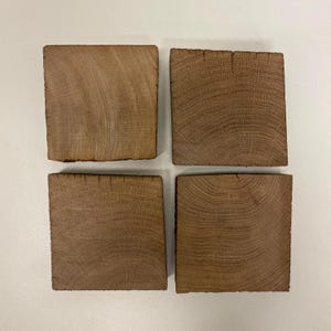 May include: A set of four square coasters made from natural brown wood. Each coaster displays a distinct grain pattern with visible growth rings and a textured, raw edge. The wood pieces are arranged in a two-by-two grid formation, showcasing the organic light and dark brown tones of the timber material.
