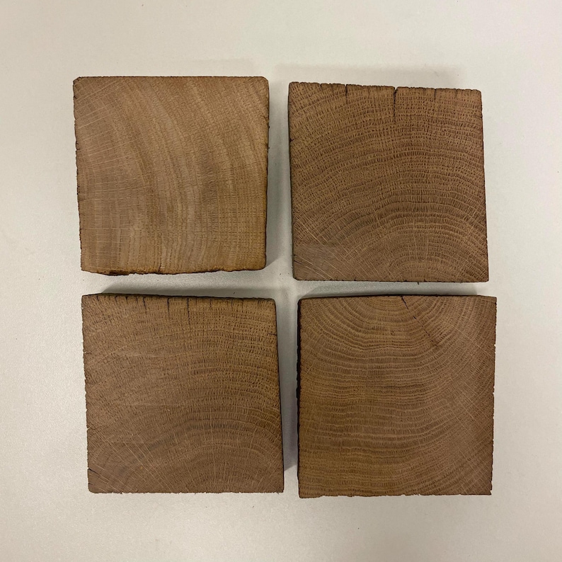 May include: A set of four square coasters made from natural brown wood. Each coaster displays a distinct grain pattern with visible growth rings and a textured, raw edge. The wood pieces are arranged in a two-by-two grid formation, showcasing the organic light and dark brown tones of the timber material.