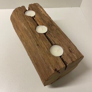 May include: A rustic wooden candle holder with three circular openings, each holding a white tea light candle. The wood has a natural, weathered appearance with visible grain and a warm brown color. The candle holder is a decorative item.