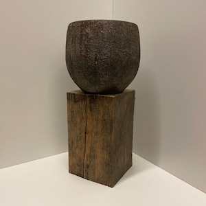 May include: A dark brown, textured, spherical cast iron vessel with a rough, weathered surface resting securely on top of a tall, rectangular, solid wooden block featuring a visible vertical grain and natural wood cracks throughout the structure.