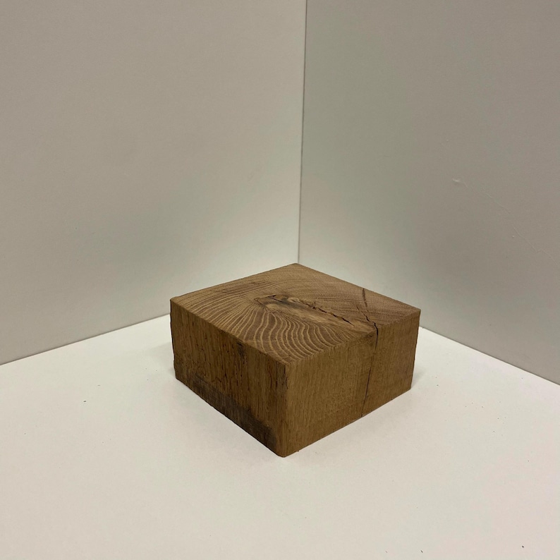 May include: A square block of natural light brown wood with a visible grain pattern and a prominent knot in the center of the top surface. The block has a smooth, flat top and straight, vertical sides, showcasing the raw texture of the timber.