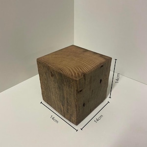 May include: A solid brown wooden cube with visible grain patterns and natural texture. The block features dimensions of 14cm by 14cm by 14cm as indicated by the measurement lines on the object. The wood has a weathered appearance with small knots.