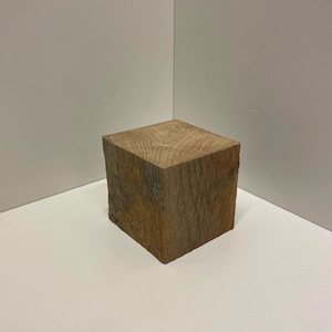 May include: A solid, cube-shaped block of natural brown wood with a visible grain pattern on the top surface. The sides of the wooden block show a rough, textured finish with vertical lines and natural variations in the wood tone and color.