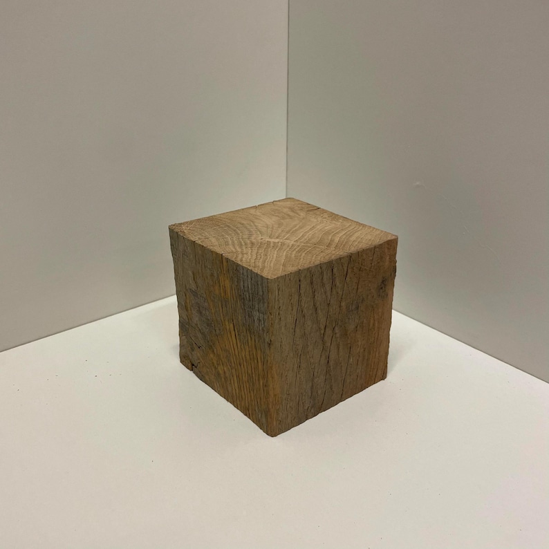 May include: A solid, cube-shaped block of natural brown wood with a visible grain pattern on the top surface. The sides of the wooden block show a rough, textured finish with vertical lines and natural variations in the wood tone and color.