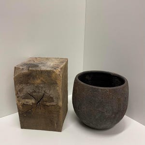 Puede incluir: Un bloque rectangular de madera marrón desgastada con vetas visibles y una grieta central en la superficie superior, junto a un cuenco de cerámica marrón oscuro redondeado con un exterior rugoso y texturizado y un acabado interior liso.