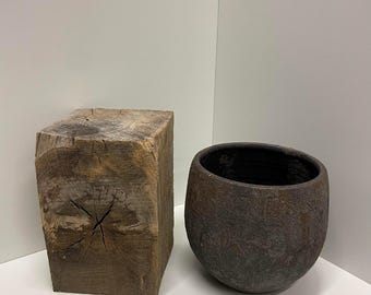 Könnte beinhalten: Ein rechteckiger Block aus verwittertem braunem Holz mit sichtbarer Maserung und einem zentralen Riss auf der Oberseite, neben einer abgerundeten, dunkelbraunen Keramikschale mit rauer, strukturierter Außenseite und glatter, dunkler Innenseite.