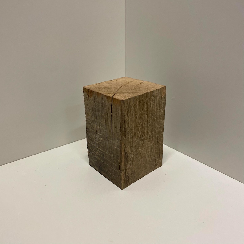 May include: A rectangular block of natural brown wood with a visible grain pattern and a rough, textured surface. The wooden block features vertical sides and a flat top, displaying organic cracks and variations in the wood tone throughout.
