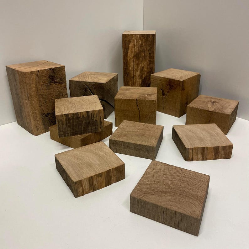 May include: A collection of twelve natural wood blocks of varying heights and square shapes, featuring visible wood grain patterns and textured bark edges in shades of light to medium brown, arranged in a cluster on a flat white surface.