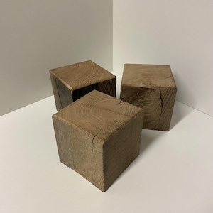 Op de afbeelding: Drie massieve houten blokken met een natuurlijke bruine afwerking, elk met een vierkant bovenoppervlak met zichtbare cirkelvormige nerfpatronen en verticale scheuren langs de zijkanten, die de ruwe textuur en organische lijnen van het hout tonen.