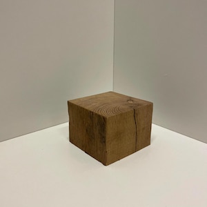 May include: A solid, square-shaped wooden block featuring a natural light brown grain pattern. The top surface displays circular wood grain rings, while one side shows a vertical crack running through the center of the textured timber surface.