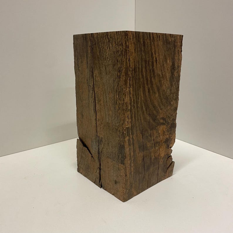 Pu&ograve; includere: Un blocco rettangolare di legno marrone scuro con una venatura visibile e una consistenza ruvida e usurata. L'oggetto presenta crepe verticali e rientranze naturali lungo i lati, mostrando un aspetto grezzo e non rifinito in ogni parte.