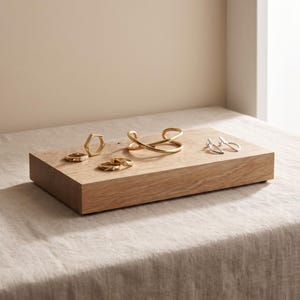 May include: A collection of gold-toned jewelry including a hexagonal ring, two simple bands, a crossover cuff bracelet, and a pair of silver-toned earrings arranged on a rectangular light-colored wooden block display stand.
