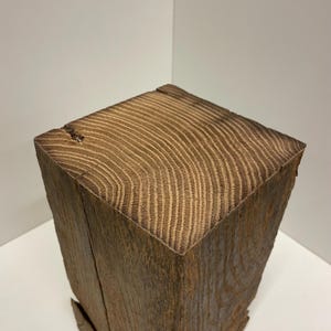 Pu&ograve; includere: Un blocco di legno di recupero marrone scuro tagliato a forma quadrata, caratterizzato da un evidente motivo a venature sulla superficie superiore. Il legno presenta anelli di crescita visibili e una consistenza ruvida e usurata sui lati verticali.
