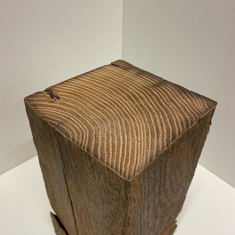 Pu&ograve; includere: Un blocco di legno di recupero marrone scuro tagliato a forma quadrata, caratterizzato da un evidente motivo a venature sulla superficie superiore. Il legno presenta anelli di crescita visibili e una consistenza ruvida e usurata sui lati verticali.